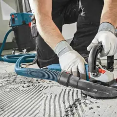 Pracownik w rękawiczkach szlifuje betonową podłogę z odkurzaczem. Sprawdź szlifowanie betonu cena za m2.