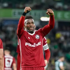 Piłkarz w czerwonej koszulce z podniesionymi pięściami na tle stadionu.