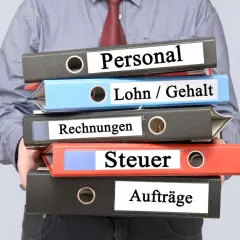 Ein Mann hält einen Stapel Ordner mit den Beschriftungen "Personal", "Lohn / Gehalt", "Rechnungen", "Steuer" und "Aufträge". Die Ordner stapeln sich und deuten auf die Komplexität der Verwaltung des zu versteuernden Einkommens hin.