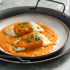 Dos lomos de bacalao a la vizcaína, cubiertos de salsa roja y perejil picado, servidos en un plato sobre una bandeja metálica.