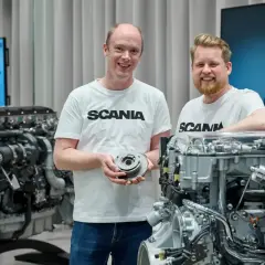Dwóch mężczyzn w koszulkach Scania prezentuje części silnika. W tle widać potężny silnik Scania V8.