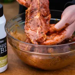 Fleisch wird mit einer würzigen Grillmarinade in einer Schüssel vermischt. Die Flasche "#Schwarzer Knobi" zeigt, wie einfach es ist, Grillmarinade selber machen.