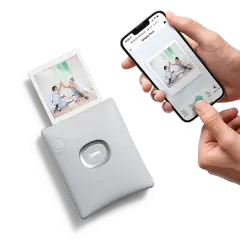 Ręce trzymają smartfon z aplikacją do drukowania zdjęć, obok drukarka instax wypuszcza gotowe zdjęcia jak z instax.