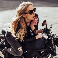 Chica rubia con gafas de sol y un perfecto outfit motero mujer, apoyada en una moto clásica.