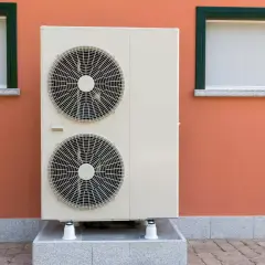 Unidad exterior de bomba de calor con dos ventiladores, instalada junto a una pared naranja con dos ventanas.