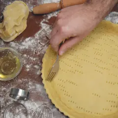 Mani che preparano una pasta frolla morbida, bucherellandola con una forchetta in una tortiera. Farina e ingredienti sparsi sul tavolo.
