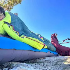 A person rests on a sleeping pad under a bright blue sky, bundled in a Nemo Disco 15 weight sleeping bag. A red backpack sits nearby.