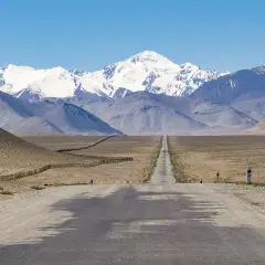 Długa, prosta droga prowadzi przez pustynny krajobraz w kierunku ośnieżonych gór. To widok na Pamir Highway, zapierający dech w piersiach szlak podróżniczy.