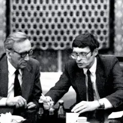Dwóch mężczyzn w garniturach przy stole, dyskutujących o historii politycznej Polski. Antoni Dudek analizuje dokumenty.