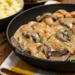 Pyszny kurczak w sosie pieczarkowym, jak u Ani gotuje, w czarnej patelni, z gałązkami rozmarynu. Obok miska z purée.