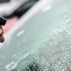 Ręka trzyma butelkę z płynem, pryskając na zamarzniętą szybę. To prosty sposób, jak zrobić odmrażacz do szyb.