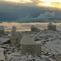 Obserwatoria astronomiczne na ośnieżonym szczycie, otoczone morzem chmur. W oddali widać złote światło zachodzącego słońca, a na pierwszym planie "la silla" - jedno z kopułowych budynków.