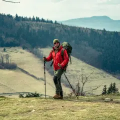 Mężczyzna w czerwonej kurtce i z plecakiem uprawia nordic walking na tle górskiego krajobrazu.
