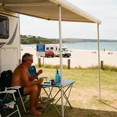 Un homme profite de son camping-car naturiste au bord de la plage, avec la mer et d'autres campeurs en arrière-plan.