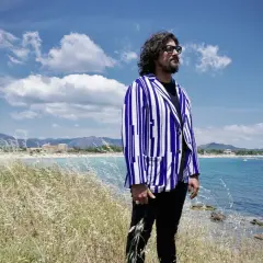 Uomo con giacca a righe blu e bianche guarda il mare in Sardegna, tra i campi di erba alta. Un'ambientazione perfetta per scoprire i **4 ristoranti Sardegna**.
