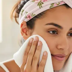 Frau mit Blumenstirnband trocknet ihr Gesicht mit einem weißen Handtuch, um Rückstände von Reinigungsmitteln wie Sodium Lauryl Sulfate zu entfernen.
