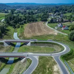 Nowoczesny węzeł drogowy na drodze krajowej 75, z mostami nad rzeką, otoczony zielonymi wzgórzami i domami.