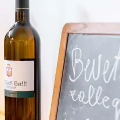 Bottiglie di vino "Est!! Est!!!" in primo piano, con una lavagna che invita a "Bevete e rallegratevi".