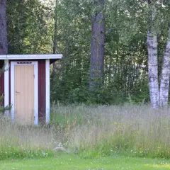 Rött utedass med gul dörr, omgivet av högt gräs och träd. Här gäller utedass regler för en trevlig vistelse.