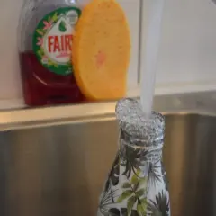 Eine Nalgene-Flasche mit Blattmuster wird in der Spüle unter fließendem Wasser gespült. Spülmittel und Schwamm sind im Hintergrund sichtbar.
