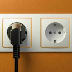 Dwa gniazdka elektryczne, jedno z wpiętą wtyczką, na pomarańczowej ścianie. Typowe gniazdka w Hiszpanii.