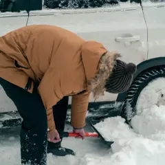 Osoba w zimowej kurtce odśnieża samochód uwięziony w zaspie śnieżnej.