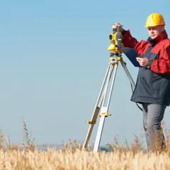 Geodeta w żółtym kasku i czerwono-granatowej kurtce pracuje z tachimetrem na polu. Zastanawiasz się, ile bierze geodeta za wytyczenie działki?