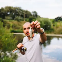 Mężczyzna trzyma małą rybkę, pokazując ją na tle jeziora. Zastanawia się, na co najlepiej bierze lin.