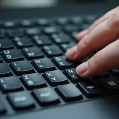 Dłoń pisze na klawiaturze, palec wskazujący naciska klawisz "C", obok widać "N" i "E". Może to być początek pisania słowa z "o umlaut na klawiaturze".