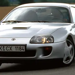 Srebrna Toyota Supra MK4 z charakterystycznym wlotem powietrza na masce, pędząca po asfaltowej drodze wśród pól.