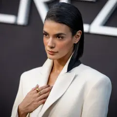Laysla de Oliveira, elegant in a white blazer, poses with a hand on her chest, adorned with a striking gold ring.