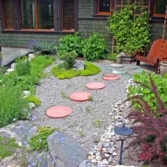 Inexpensive stepping stone walkway ideas: red circular pavers create a path through a gravel garden with lush greenery and a wooden chair.