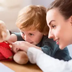 Uśmiechnięta opiekunka bawi się z dzieckiem pluszowym misiem. Zastanawiasz się, ile bierze opiekunka do dzieci za taką troskę?