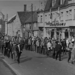 Demonstration am 7. Oktober, Tag der Republik DDR. Menschen marschieren durch eine Straße mit Geschäften, Fahnen wehen.
