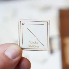 Main tenue par un pouce, une petite carte en bois gravée indique "Douche 80x80cm", une dimension pour une future douche.