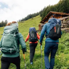 Tres excursionistas con mochilas y bastones suben por una ladera verde hacia una cabaña de madera. Saben qué llevar a la montaña.