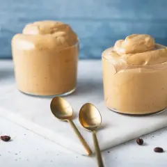 Due bicchieri di cremosa schiuma di caffè, perfetta per la tua ricetta crema di caffè senza panna. Due cucchiaini dorati e chicchi di caffè completano la scena.