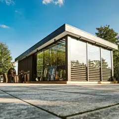 Nowoczesna zabudowa altany przed wiatrem, z przeszklonymi ścianami i drewnianymi elementami, na tarasie z kamienną nawierzchnią.