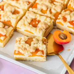 Saftiger aprikosenkuchen mit Streuseln, auf einem Blech arrangiert. Ein Stück wird mit einem goldenen Tortenheber herausgehoben.