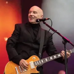 Łysy mężczyzna w czarnym garniturze gra na gitarze elektrycznej, uśmiechając się. To jest jego chwila, bez żalu.