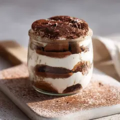 Warstwowe tiramisu z ricotty w słoiku, posypane kakao. Delikatny deser na desce, skąpany w słońcu.