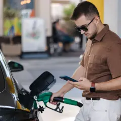 Un homme fait le plein de sa voiture à une **téléphone station essence**, consultant son smartphone.