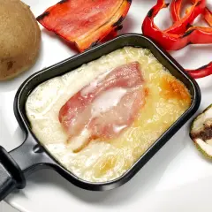 Un poêlon de raclette avec fromage fondu et lardons, accompagné d'une pomme de terre, de poivrons rouges grillés et de courgettes. Une recette de sauce raclette facile et gourmande.