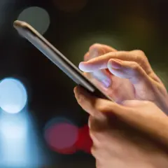 Dłoń trzyma smartfon, palec wskazujący dotyka ekranu, być może w trakcie odzyskiwania zdjęć z telefonu. Rozmyte światła w tle sugerują noc.