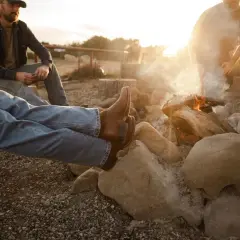 Two men relax by a campfire. One wears brown boots, and the other, a cap. The sun sets, casting a warm glow.