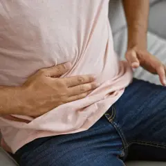 Mężczyzna siedzi na kanapie, trzymając się za brzuch. Odczuwa silny ból jelit, który utrudnia mu codzienne funkcjonowanie.