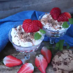 Två glas med krämig dessert toppad med jordgubbar och smulor. Bredvid ligger halverade jordgubbar och chokladkakor. En härlig sommardessert med jordgubbar och glass.