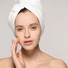 Kobieta z ręcznikiem na głowie delikatnie masuje twarz pokrytą peelingiem. Poznaj, jak stosować peeling do twarzy dla promiennej cery.