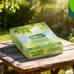 Pudełko z napisem "Vegano Dobroczynek Kalifornijski" na drewnianym stole w ogrodzie pełnym kwiatów.