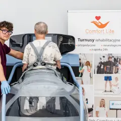 Rehabilitacja po operacji kręgosłupa: pacjent w specjalistycznym urządzeniu, asystuje mu terapeuta. Na banerze informacje o turnusach.
